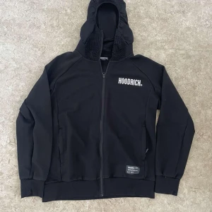 Svart hoodie från Hoodrich med tryck - Svart hoodie från Hoodrich med vit logga på bröstet och coolt tryck på ryggen. Tröjan har hel dragkedja, huva med mesh-detaljer och långa ärmar. Perfekt för dig som gillar streetwear och vill ha en stilren look.