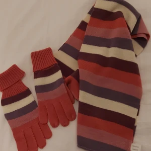 Randig halsduk och vantar, i rosa och lila toner med lite glitter - Halsduken är mjuk och har en klassisk rektangulär form. Perfekt för att hålla värmen under kyliga dagar. Matchande vantar ingår!