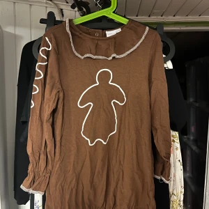 Pepparkaksklänning från Mywear Young - Brun kortklänning från Mywear Young med vit pepparkaksgubbe framtill och vita vågiga detaljer på ärmarna. Klänningen har långa ärmar, volangkant vid halsen och är gjord i mjuk bomull. I den här beställningen så får du också med ett diadem med pepparkakor och en hat/huvudbonad som matchar med klänningen!  Klänningen stängs med hjälp av knappar och huvudbonaden har ett resorband så att det är enkelt att ha på sig den! Storlek 122. Perfekt för alla julens tillfällen! Pris går att diskutera! 💕