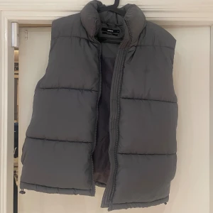 Grå dunväst från BikBok - Säljer en grå dunväst från BikBok i storlek S. Västen har en hög krage, breda quiltade paneler och dragkedja framtill. 