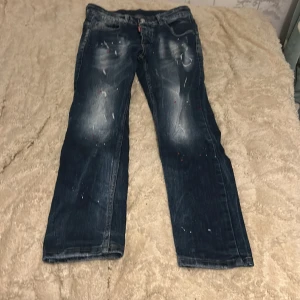 Dsquared2 blå jeans med färgstänk - Säljer ett par blå jeans från Dsquared2 med coola slitningar och färgstänk i rött och vitt. Jeansen har en rak passform, klassiska fem fickor och en röd Dsquared2-label bak i midjan. Materialet är jeans i bomull och de har en snygg tvättad look.