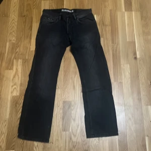 Svarta jeans från Blend Denim, stl 31 - Svarta jeans från Blend Denim med raka ben och klassisk femficksdesign. Jeansen har en mörk tvätt och detaljerad bakficka med Blend-logga samt ett mörkrött läderpatch bak i midjan. Perfekta för dig som gillar en enkel och stilren look.