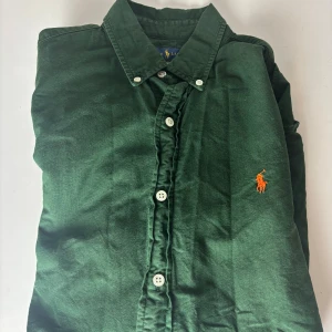 Grön skjorta från Ralph Lauren M - Snygg grön skjorta från Ralph Lauren i slim fit med klassisk krage och vita knappar. Broderad orange logga på bröstet ger en stilren touch. Skjortan är långärmad och tillverkad i mjuk bomull, perfekt för dig som gillar en clean och tidlös look.