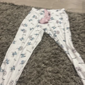 Stitch-mönstrade mjukisbyxor med snörning - Supermysiga vita mjukisbyxor med Stitch och Angel-motiv i blått och rosa. Byxorna har en rosa snörning i midjan och är perfekta för chill dagar hemma. Tillverkade i mjukt bomullsmaterial för extra komfort.