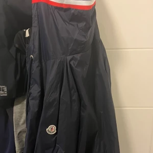 Mörkblå vindjacka från Moncler - Snygg mörkblå vindjacka från Moncler med klassisk logga på ärmen. Jackan har en röd och vit rand upptill, dragsko i midjan och praktiska fickor med knapp. Perfekt för dig som vill ha en stilren och sportig look.