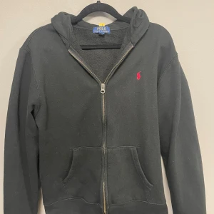 Svart zip hoodie från Polo Ralph Lauren  - Svart hoodie från Polo Ralph Lauren i storlek M med dragkedja framtill och klassisk huva. Tröjan har en röd broderad logga på bröstet och två fickor framtill. Perfekt för chill dagar och enkel att matcha med jeans eller joggers.