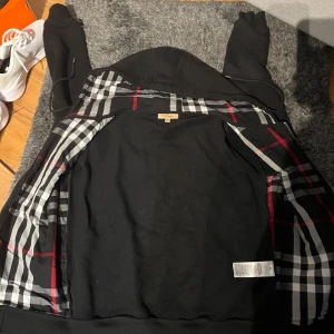 Svart hoodiejacka från Burberry SP - Svart hoodiejacka från Burberry med klassiskt rutigt foder i svart, vitt och rött. Jackan har dragkedja framtill, snörning i huvan och två fickor. Materialet är mjuk bomull med lite elastan för extra komfort. Perfekt för dig som gillar stilren design med lyxig touch.