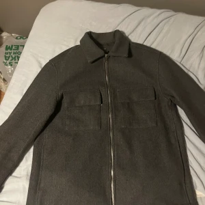 Mörkgrå overshirt med dragkedja - Snygg mörkgrå overshirt från H&M med klassisk krage och två stora bröstfickor. Jackan har en rak passform och stängs med en silverfärgad dragkedja framtill. Perfekt att slänga på över en hoodie eller t-shirt för en chill look. Öppen för bud