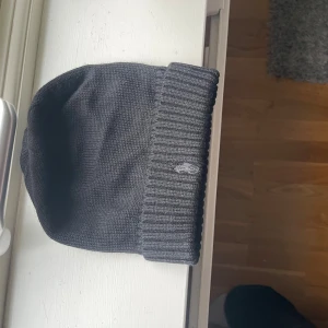 Mörkgrå mössa från Polo Ralph Lauren - Snygg mörkgrå mössa från Polo Ralph Lauren i 100% ull. Klassisk ribbstickad kant och diskret broderad logga framtill. Perfekt accessoar för kalla dagar och enkel att matcha med olika outfits. Nypris 700kr