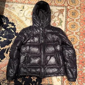 Svart pufferjacka från H&M med huva - Svart pufferjacka från H&M med glansig yta och stor huva. Jackan har dragkedja framtill, två sidofickor med dragkedja och elastiska muddar vid ärmslut. Perfekt för kalla dagar och har en schysst, lite oversized passform.