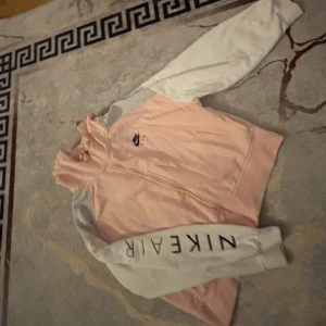 Nike Air rosa och vit vindjacka - Säljer en snygg Nike Air vindjacka i ljusrosa med vita ärmar och svart logga på bröstet. Jackan har hel dragkedja framtill och tryckt Nike Air-text på ena ärmen. Perfekt för dig som gillar sportig stil och streetwear.