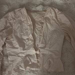 Ljusbeige blus med knappdetalj - Säljer en ljusbeige blus med lång ärm och en snygg v-ringning. Blusen har en stor rund knapp framtill som stängning och är tillverkad i ett tunt, mjukt material som ger en skön känsla. Perfekt för dig som gillar stilrena och enkla plagg med en twist.