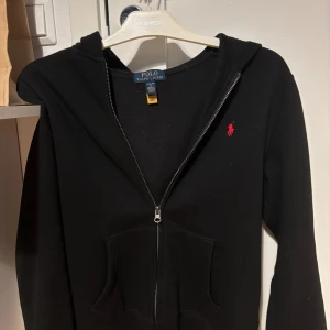 Svart hoodie från Polo Ralph Lauren - Svart hoodie från Polo Ralph Lauren med dragkedja och huva. Klassisk design med röd broderad logga på bröstet och stora fickor framtill. Perfekt för en chill och stilren look.