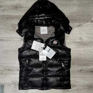 Svart dunväst  Moncler med huva - Svart Moncler Vest i storlek M. Jackan har 3 fickor 2 framsida och en inuti (QR-Kod Skan och autencitet papper finns inuti lilla fickan). Helt ny med alla taggar kvar och passar i princip med alla färger. 
