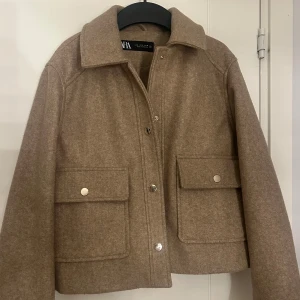 Beige jacka från Zara - Snygg beige jacka från Zara i storlek S. Jackan har två stora fickor framtill med guldiga knappar, klassisk krage och rak passform. Materialet är mjukt och känns lite ulligt, perfekt för lager-på-lager. Enkel och clean look som funkar till många stilar.