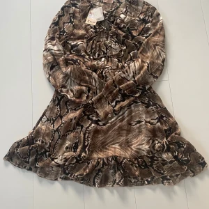 Brun klänning Buch Copenhagen  - Säljer en kort klänning från Buch Copenhagen i storlek M med coolt mönster, bruna och beige toner. Klänningen har långa ärmar, snörning framtill och volangkant nedtill. Materialet är glansigt och känns lyxigt mot huden. Perfekt för dig som gillar att sticka ut. Nypris: 700 sek
