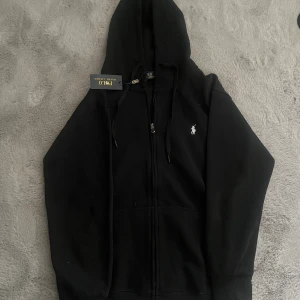 Svart hoodie från Polo Ralph Lauren - Svart hoodie från Polo Ralph Lauren med dragkedja framtill, huva med snörning och klassisk vit logga broderad på bröstet. Tröjan har ribbade muddar och känguruficka. Perfekt för en chill och stilren look. Passar till s 