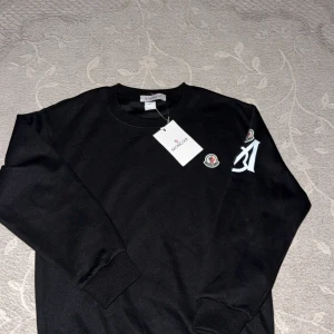 Svart Moncler sweatshirt med logga - Svart sweatshirt från Moncler med rund halsringning, broderad logga på bröstet och vit tryckt detalj på ärmen. Tröjan har långa ärmar och ribbade muddar vid ärmslut och nederkant. Perfekt för en stilren och trendig look.