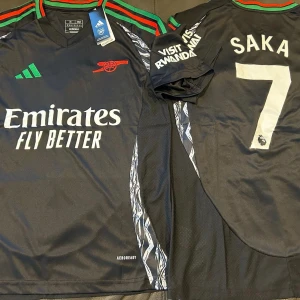Arsenal Saka 7 svart bortatröja Adidas - Arsenal bortatröja med Saka 7-tryck, svart bas med vita detaljer och gröna samt röda ränder i kragen. Adidas-logga i grönt, klubbmärke i rött och sponsortryck framtill. Tillverkad i lätt och ventilerande polyester med mönstrade inslag längs sidorna.