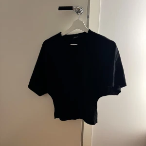 Svart topp med vida ärmar - En svart topp medvida, korta ärmar. Toppen har en enkel och stilren design med rund halsringning och är tillverkad i mjukt bomullstyg. Perfekt för en avslappnad och trendig look.