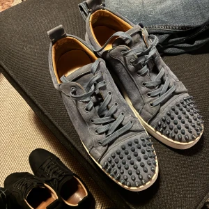 Christian Louboutin grå mocka sneakers - Säljer ett par grå sneakers från Christian Louboutin i mjuk mocka med ikoniska röda sulor. Skorna har snörning och coola nitar på tån för extra edge. Insidan är beige och sulan är vit med röd undersida. Perfekt för dig som vill sticka ut med lyxig streetstyle.