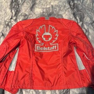 Röd vindjacka från Belstaff - Snygg röd vindjacka från Belstaff med stora loggan broderad på ryggen och flera praktiska fickor framtill. Jackan har silvriga detaljer, dragkedja och tryckknappar, samt meshfoder på insidan. 