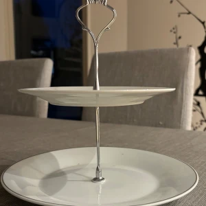 Kakfat - Tvåvånings kakfat i porslin med metallhandtag. Fatet är vitt med diskret silverkant och är i mycket gott skick utan repor eller skador. Perfekt för servering av kakor, bakverk eller frukt vid festliga tillfällen.