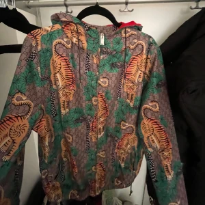 Gucci vindjacka med tigerprint - Gucci vindjacka med ikoniskt tigerprint i orange och svart på grå bakgrund med gröna blad. Jackan har huva, dragkedja framtill och röd insida. Snygg och unik design som sticker ut, perfekt för dig som vill ha något extra.