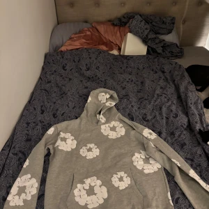 Grå hoodie från Denim Tears med blommor - Säljer en grå hoodie från Denim Tears med vita blommiga tryck över hela tröjan. Hoodien har huva och är tillverkad i USA. Perfekt för dig som vill ha en unik och snygg streetwear-look.