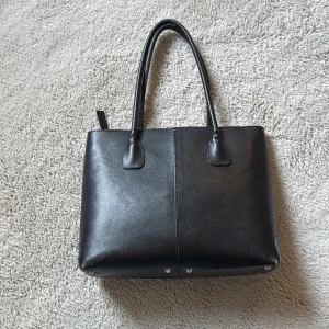 Svart handväska från Lottusse i skinn - Stilren svart handväska från Loxwood i äkta skinn med två långa handtag. Väskan har en klassisk fyrkantig form och stängs med dragkedja. Perfekt storlek för att få plats med allt du behöver och passar till många olika outfits.