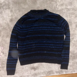 Glittrig svart-blå randig tröja - Säljer en svart tröja med blå glittriga ränder. Tröjan har lång ärm, rund hals och är stickad i ett glittrigt material som ger en snygg effekt. Perfekt för dig som vill sticka ut lite extra. Passar dig som gillar en normal passform.