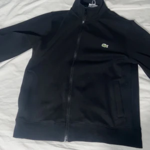 Svart ziptröja från Lacoste - Svart ziptröja från Lacoste med klassisk krokodillogga på bröstet. Tröjan har hög krage, hel dragkedja och två fickor framtill. Tillverkad i mjuk bomullsmix, perfekt för lager-på-lager. Enkel och stilren design som funkar året runt.