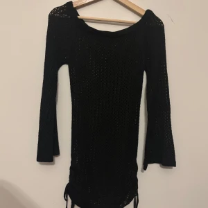 Svart stickad strandklänning - Svart stickad strandklänning med lång utsvängd ärm och snyggt hålmönster. Klänningen har snörning i sidorna nertill för en extra detalj. Perfekt för dig som gillar en enkel men ändå trendig look till stranden.