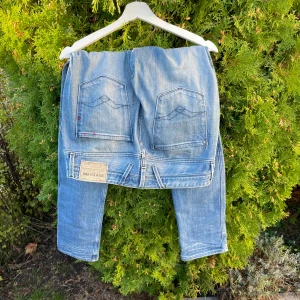 Replay Anbass ljusblå jeans, stl 32 - Säljer ett par Replay Anbass jeans i ljusblå tvätt med snygga slitningar och klassiska femficksdetaljer. Jeansen har röd brodyr på insidan, normal passform och raka ben. Märkespatch bak och coola sömmar på bakfickorna. Perfekta för en avslappnad streetstil.