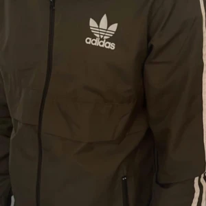 Svart vindjacka från Adidas med huva - Svart vindjacka från Adidas med klassiska vita ränder på ärmarna och huvan. Jackan har dragkedja framtill, Adidas-logga på bröstet och praktiska fickor. Perfekt för dig som gillar sportig och clean stil.