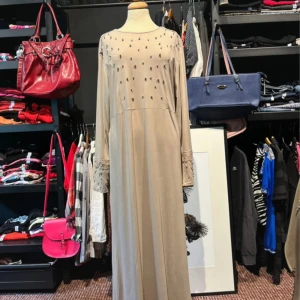 Beige långklänning från Vogue  - Beige långklänning från Vogue i storlek XL, tillverkad i mjuk polyester. Klänningen har lång ärm med spetsdetaljer och små pärlor framtill. Den har ett bälte i midjan och en elegant, rak siluett som ger en stilren look.