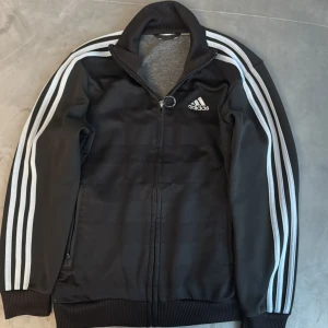 Svart Adidas track jacket med dragkedja - Svart Adidas track jacket med klassiska vita ränder längs ärmarna och broderad logga på bröstet. Hög ribbad krage, hel dragkedja framtill och ribbade muddar. Tillverkad i glansig polyester, perfekt för en sportig och stilren look.