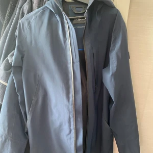 Mörkblå regnjacka från Sail Racing - Snygg mörkblå regnjacka från Sail Racing med huva och diskret logga på ärmen. Jackan har dragkedja framtill, minimalistisk design och är tillverkad i ett slätt, vattenavvisande material. Perfekt för blöta dagar och stilren look. Använd men i bra skick 