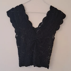 Svart spetsblus från Zara - Supersnygg svart blus från Zara i spets med v-ringning och breda axelband. Blusen har vågiga kanter och är croppad, perfekt att styla med höga jeans eller kjol. Materialet är genombruten spets som ger en cool och feminin look.
