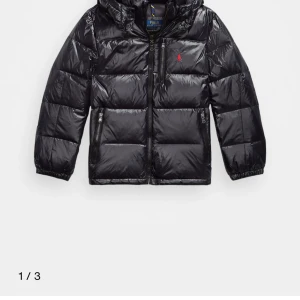 Svart pufferjacka från Polo Ralph Lauren - Svart pufferjacka från Polo Ralph Lauren med glansig finish, huva och dragkedjor på fickor och framtill. Jackan har en röd broderad logga på bröstet och justerbar dragsko nedtill. Perfekt för kalla dagar och riktigt snygg streetstil.