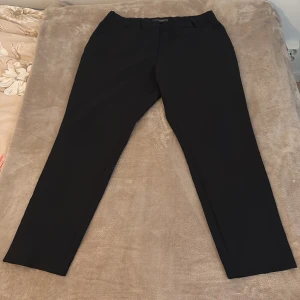 Svarta kostymbyxor från Primark - Svarta kostymbyxor från Primark med klassisk rak passform och bälteshällor. Byxorna har diskret dragkedja och knapp framtill samt röda detaljer på insidan av linningen. Perfekta för en clean och stilren look.