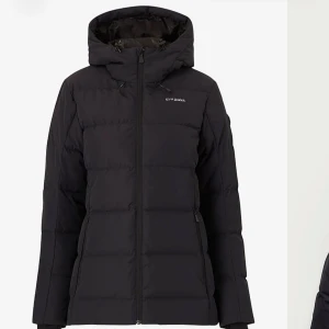 Svart dunjacka från ÁHKKÁ - Svart dunjacka från ÁHKKÁ med huva och dragkedja framtill. Jackan är quiltad med horisontella sömmar och har diskret logga på bröstet. Perfekt för kalla dagar med sin långa passform och praktiska fickor. Loggan på bröstet kan se annorlunda ut på de exemplaret jag säljer, men likt.