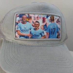 Grå meshkeps med fotbollstryck - Säljer en unik grå keps med mesh på sidorna och baksidan samt justerbar snapback. Framtill finns ett färgstarkt tryck med fotbollsspelare i ljusblå tröjor. Kepsen har böjd skärm och är tillverkad i syntetmaterial och mesh för extra ventilation.