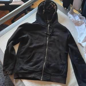 Svart Burberry hoodie med dragkedja - Svart hoodie från Burberry med klassiskt rutigt foder i huvan och innerkant. Broderad logga på bröstet, hel dragkedja och känguruficka framtill. Mjuk bomullsblandning och snörning i huvan. Perfekt för en chill och stilren look.