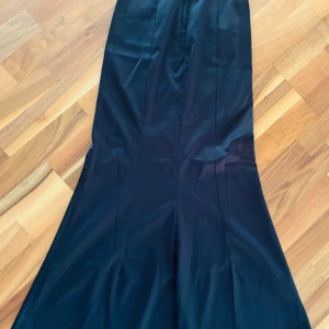 Svart långklänning med utsvängd kjol - En elegant festlig svart långkjol med figurnära passform upptill och utsvängd kjol nertill. Klänningen har en stilren och tidlös design utan synliga detaljer eller mönster, perfekt för dig som gillar en klassisk look.