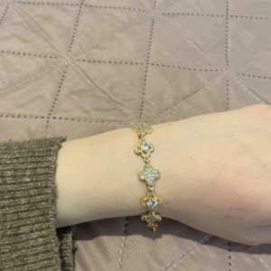 Guldigt armband med glittriga detaljer - Snyggt armband i guld med flera fyrklöverformade länkar som är täckta av glittrande stenar. Perfekt för dig som vill ha något extra på handleden och gillar bling. Armbandet har en elegant och lyxig vibe.