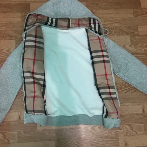 Grå Burberry hoodie med dragkedja - Grå hoodie från Burberry med klassiskt rutigt foder i huvan och längs insidan. Använder den inte längre har bara använt den några gånger. Men annars e den som vanligt ny och inga skador eller så och vill sälja den eftersom jag inte rockar den längre. Storlek M fråga gärna om ni undrar något eller är intresserade😁!