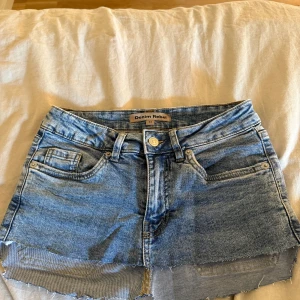 Blå jeanskjol från Denim Rebel - Snygg blå jeanskjol från Denim Rebel med rå kant nertill och klassiska femficksdetaljer. Kjolen har silverfärgade nitar på bakfickorna och en cool, avslappnad look. Perfekt för dig som gillar denim och vill ha en kort kjol med attityd.