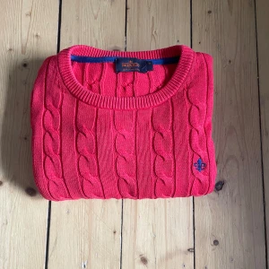 Röd kabelstickad tröja från Morris - Säljer en röd kabelstickad tröja från Morris med rund halsringning och broderad logga på bröstet. Tröjan är i bomull och har långa ärmar samt ribbade muddar för en klassisk look. Perfekt för dig som gillar stilrena och färgstarka plagg.