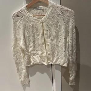 Vit stickad kofta från NA-KD - Superfin vit stickad kofta från NA-KD med snyggt hålmönster och rund halsringning. Koftan har långa ärmar, ribbade muddar och knäpps med pärlemorskimrande knappar framtill. Perfekt att slänga över en topp för en chill look.